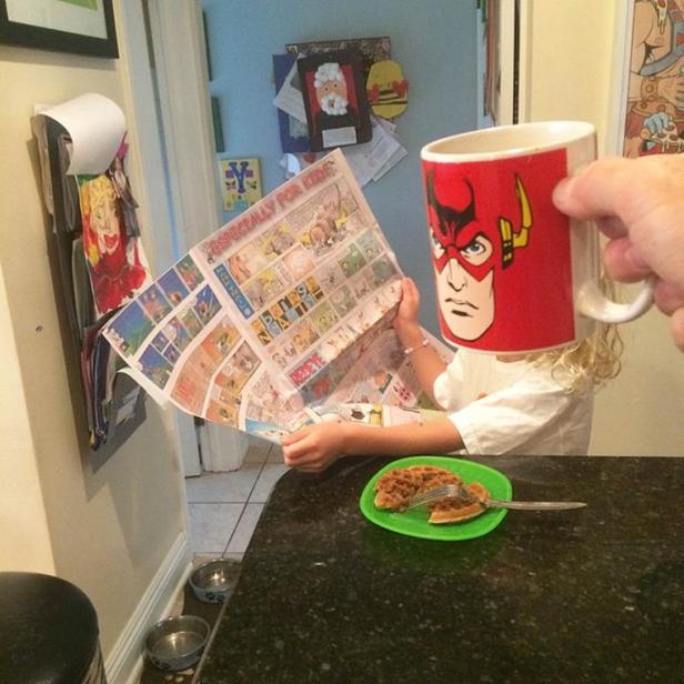 Ein Kind liest eine Zeitung hinter einer Tasse mit dem Gesicht von Flash, während es Waffeln isst.
