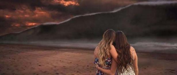 Zwei Frauen umarmen sich am Strand und blicken auf eine dunkle Wolkenwand am Horizont.