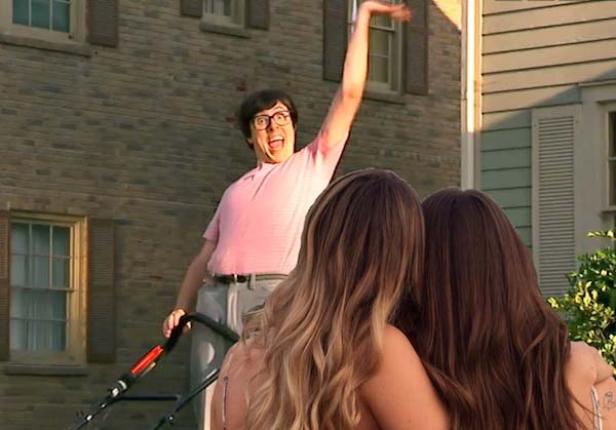 Ein Mann mit Brille und rosa Hemd steht mit einem Rasenmäher vor zwei Frauen und winkt.