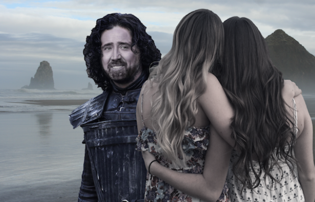 Zwei Frauen umarmen sich am Strand, während im Hintergrund eine Figur in Rüstung steht.