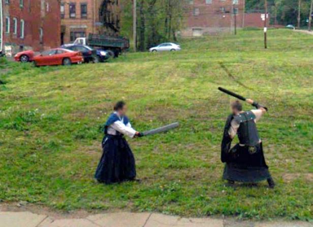 Zwei Personen in traditioneller Kleidung kämpfen mit Schwertern auf einer Wiese.