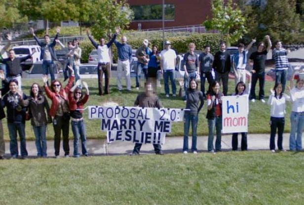Eine Gruppe von Menschen hält Schilder mit der Aufschrift „Proposal 2.0: Marry me Leslie!!“ und „Hi Mom“.