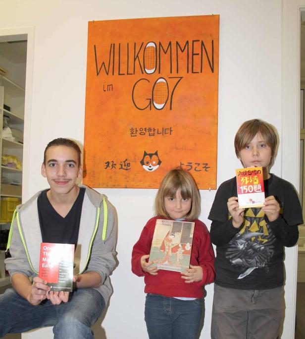 Drei Kinder stehen vor einem Poster mit der Aufschrift „Willkommen im GO7“ und halten Bücher in den Händen.