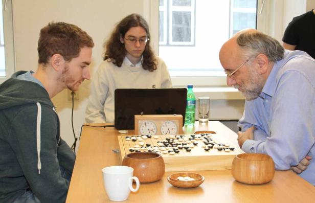 Drei Personen sitzen an einem Tisch und spielen Go.