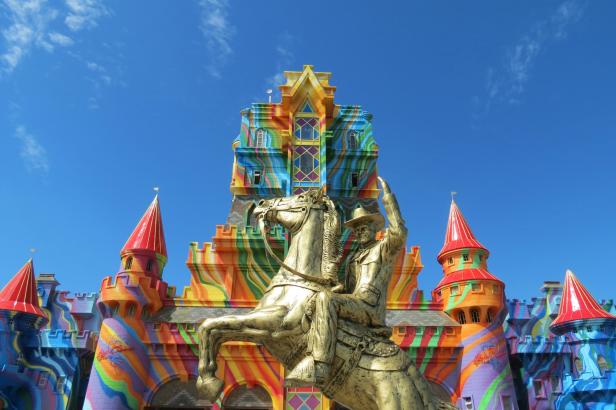 Eine goldene Reiterstatue vor einem bunten Schloss unter blauem Himmel.