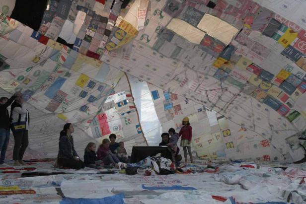 Eine Gruppe von Menschen sitzt in einem Zelt, das aus Einkaufstüten gefertigt ist.