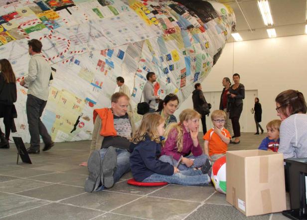 Eine Gruppe von Menschen, darunter Kinder, sitzt vor einer großen, patchworkartigen Skulptur.