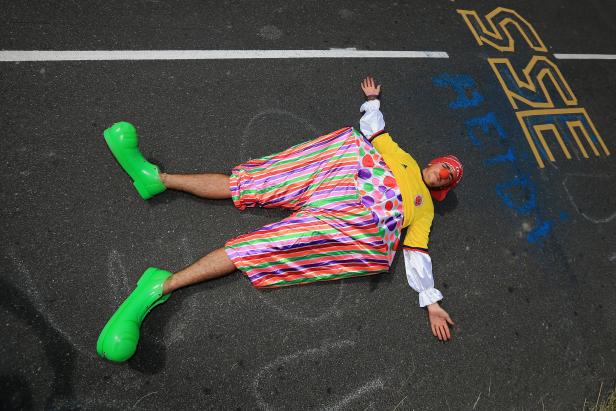 Ein Clown mit roten Haaren und übergroßen grünen Schuhen liegt auf dem Asphalt.