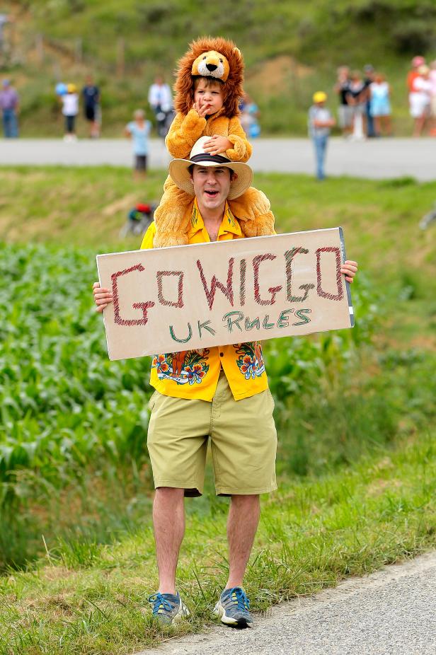 Ein Mann mit einem Kind im Löwenkostüm auf den Schultern hält ein Schild mit der Aufschrift „Go Wiggo UK Rules“.