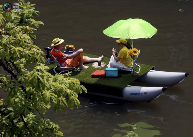 Ein Paar entspannt sich auf einem kleinen, selbstgebauten Floß mit Rasen und Sonnenschirm auf dem Wasser.