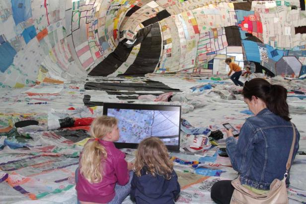 Zwei Mädchen schauen einen Fernseher in einer Installation aus Plastiktüten. Eine Frau benutzt ein Handy.
