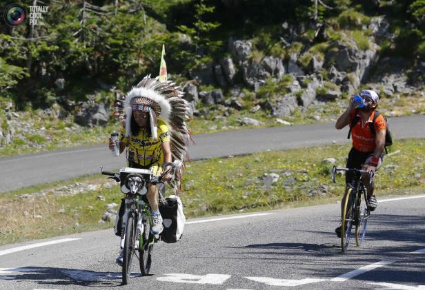 Ein Radfahrer mit Federschmuck fährt eine Bergstraße entlang, ein anderer trinkt im Hintergrund.