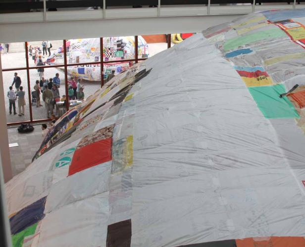 Eine große, patchworkartige Kuppel in einem Ausstellungsraum mit Besuchern im Hintergrund.