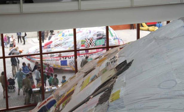 Eine große, patchworkartige Skulptur wird von Besuchern in einem Innenraum betrachtet.