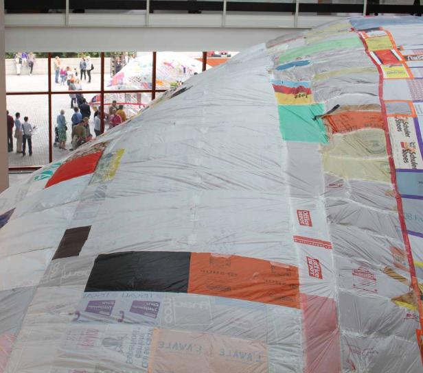 Eine große Kuppel, bedeckt mit recycelten Plastiktüten und Logos, steht in einem Gebäude.