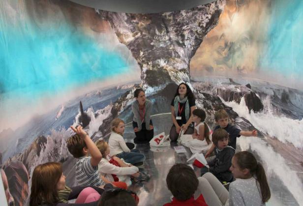 Eine Gruppe Kinder sitzt mit zwei Betreuerinnen vor einem großen Wandbild mit einer Meereslandschaft.