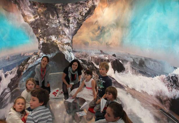 Eine Gruppe von Kindern und zwei Frauen sitzen vor einem großen Wandbild mit einer Küstenlandschaft.