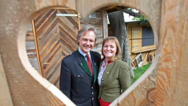 Ein Paar in Tracht steht vor einer Holzhütte, gerahmt von einem Holzbogen in Herzform.