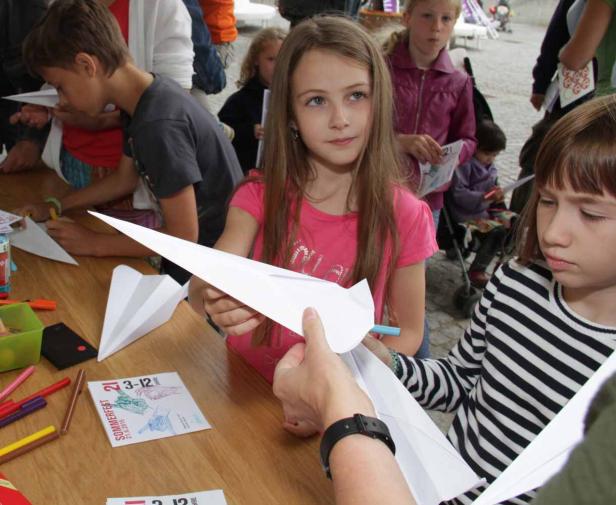 Kinder basteln Papierflieger an einem Tisch mit Stiften und Flyern.