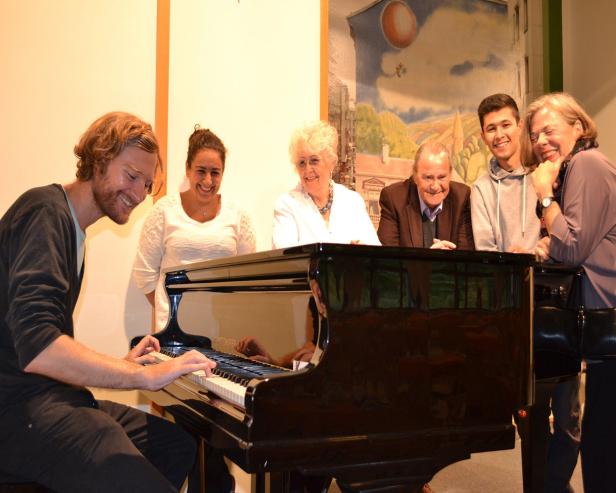 Ein junger Mann spielt Klavier, während eine Gruppe von Menschen ihm aufmerksam zuhört.