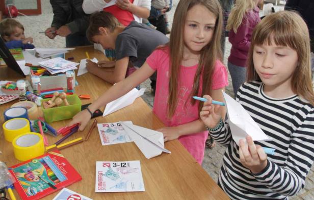 Kinder basteln Papierflieger an einem Tisch mit Stiften und Flyern.