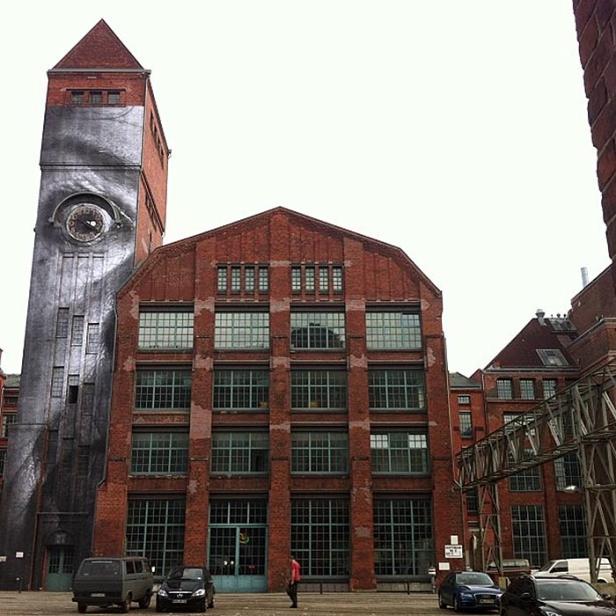 Ein hoher Backsteinturm mit einem riesigen aufgemalten Auge neben einem Backsteingebäude.