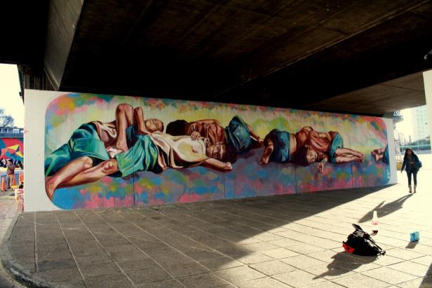 Ein Wandgemälde unter einer Brücke zeigt schlafende Menschen auf farbenfrohem Hintergrund.