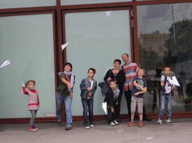Eine Gruppe von Kindern und Erwachsenen wirft Papierflieger vor einem Gebäude.