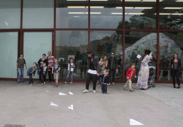 Eine Gruppe von Menschen, darunter viele Kinder, steht vor einem Gebäude und wirft Papierflieger.