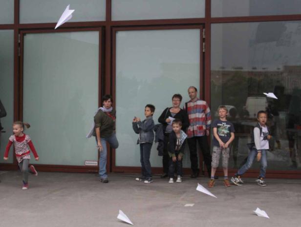 Eine Gruppe von Kindern und Erwachsenen wirft Papierflieger vor einem Gebäude.