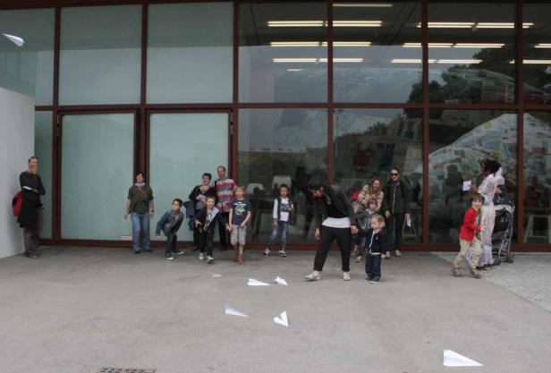 Eine Gruppe von Kindern und Erwachsenen lässt Papierflieger vor einem Gebäude steigen.