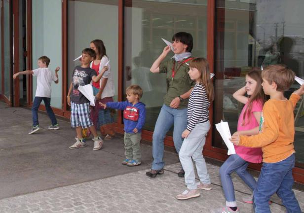 Eine Gruppe von Kindern und eine Frau spielen mit Papierfliegern vor einem Gebäude.