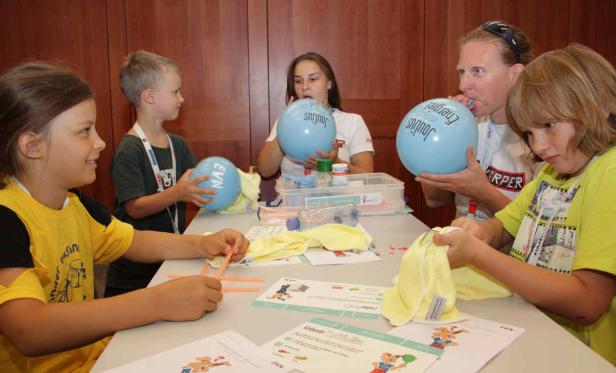 Kinder und Erwachsene sitzen an einem Tisch und basteln mit hellblauen Luftballons und gelben Tüchern.