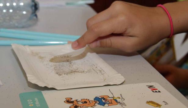 Ein Kind hantiert mit einem Papierstreifen und Samen auf einem Pappteller.