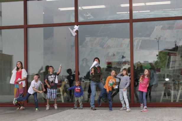 Eine Gruppe von Kindern wirft Papierflieger vor einem großen Fenster.
