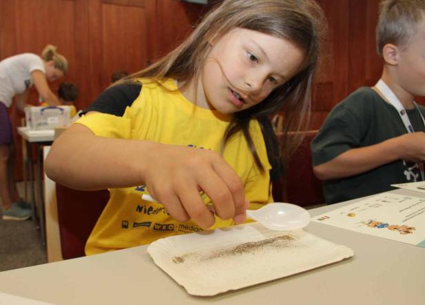 Ein Mädchen experimentiert mit einem Plastiklöffel und Sand auf einem Pappteller.