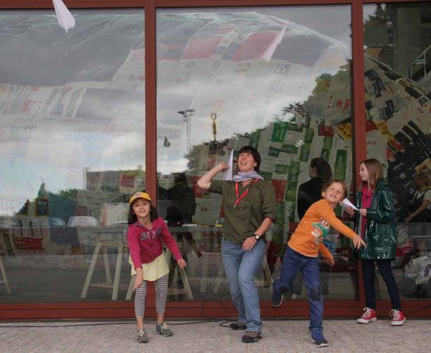 Eine Frau und drei Kinder werfen Papierflieger vor einem großen Fenster.