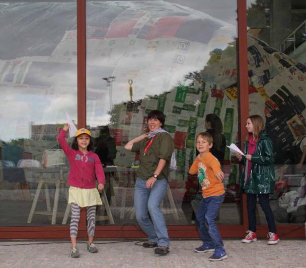 Eine Gruppe von Kindern und eine Frau posieren vor einem Gebäude, dessen Fassade mit Einkaufstüten verziert ist.