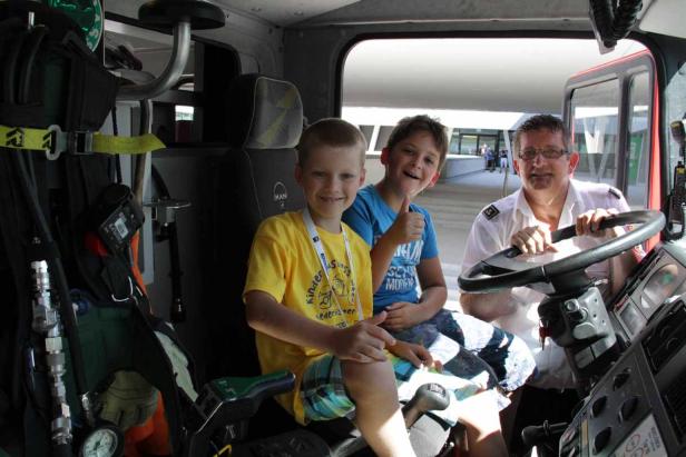 Zwei Jungen sitzen mit einem Feuerwehrmann im Führerhaus eines Feuerwehrautos.