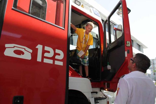 Ein Junge klettert in ein rotes Feuerwehrauto, während ein Feuerwehrmann zusieht.