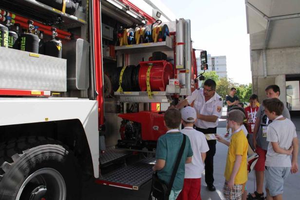 Ein Feuerwehrmann erklärt einer Gruppe von Kindern die Ausrüstung eines Feuerwehrwagens.
