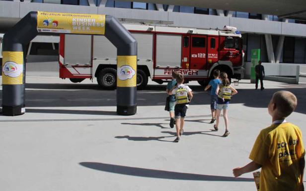 Kinder laufen auf ein Feuerwehrauto zu, das unter einem Torbogen der „Kinder Business Week“ steht.