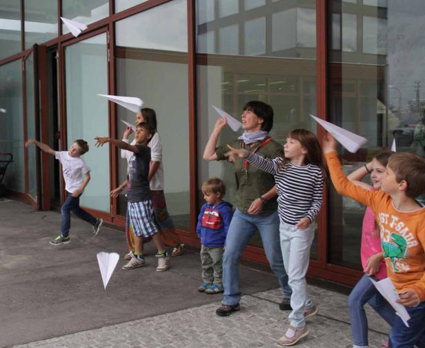 Eine Gruppe von Kindern und eine Frau werfen Papierflugzeuge vor einem Gebäude.