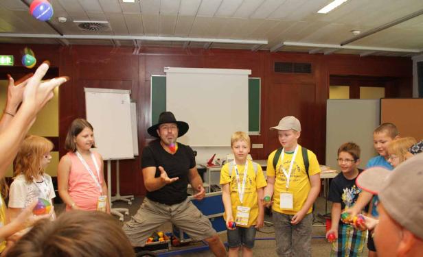 Ein Jongleur mit Hut zeigt Kindern Kunststücke mit Bällen.