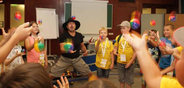 Ein Jongleur mit Hut zeigt Kindern, wie man mit bunten Bällen jongliert.