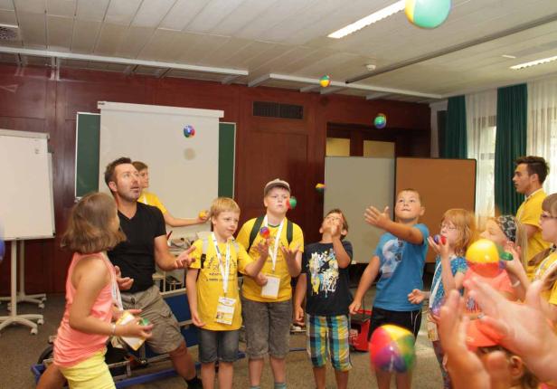 Eine Gruppe Kinder und ein Mann üben das Jonglieren mit bunten Bällen in einem Raum.