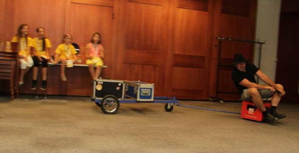 Ein Mann fährt auf einer Bierkiste, gezogen von einem Wagen mit Koffern; im Hintergrund sitzen Kinder.