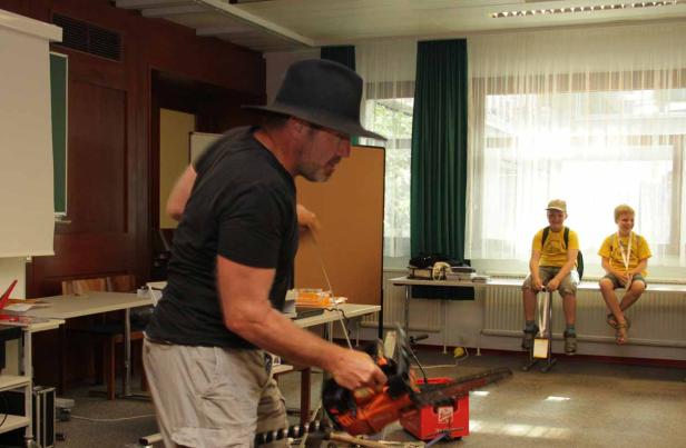 Ein Mann mit Hut hantiert mit einer Kettensäge, während zwei Jungen im Hintergrund zusehen.