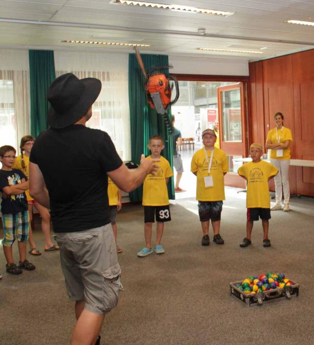 Ein Mann mit Hut jongliert mit einer Kettensäge vor einer Gruppe von Kindern.