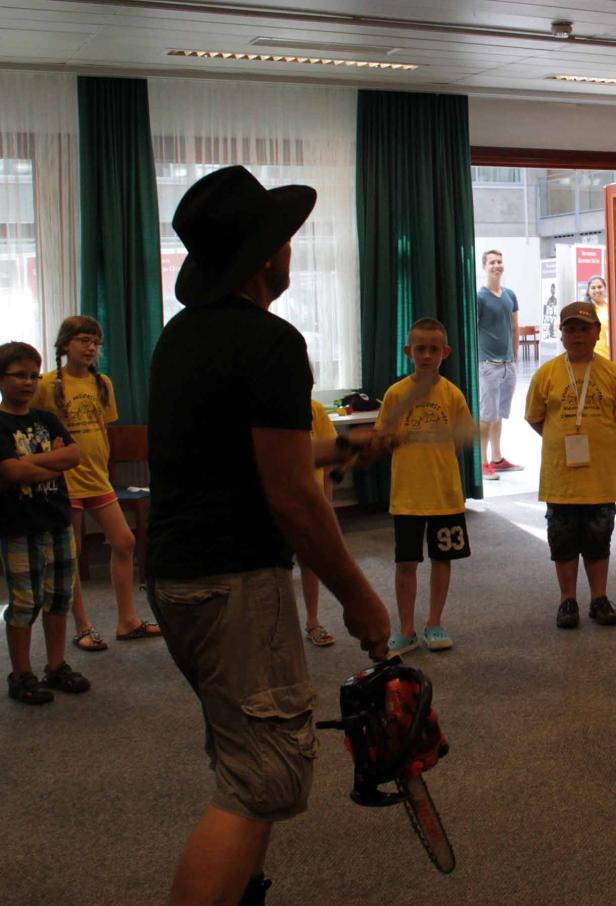 Ein Mann mit Hut hält eine Kettensäge vor einer Gruppe von Kindern.